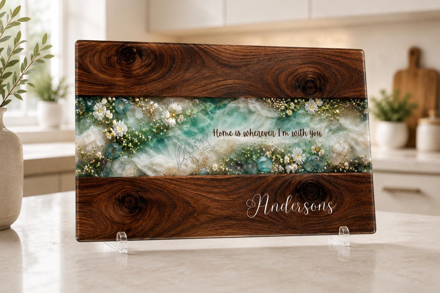 Personalised glass chopping board with dark wood effect, turquoise resin-style floral centre, white wildflowers and custom Andersons family name, displayed upright in a bright modern kitchen.