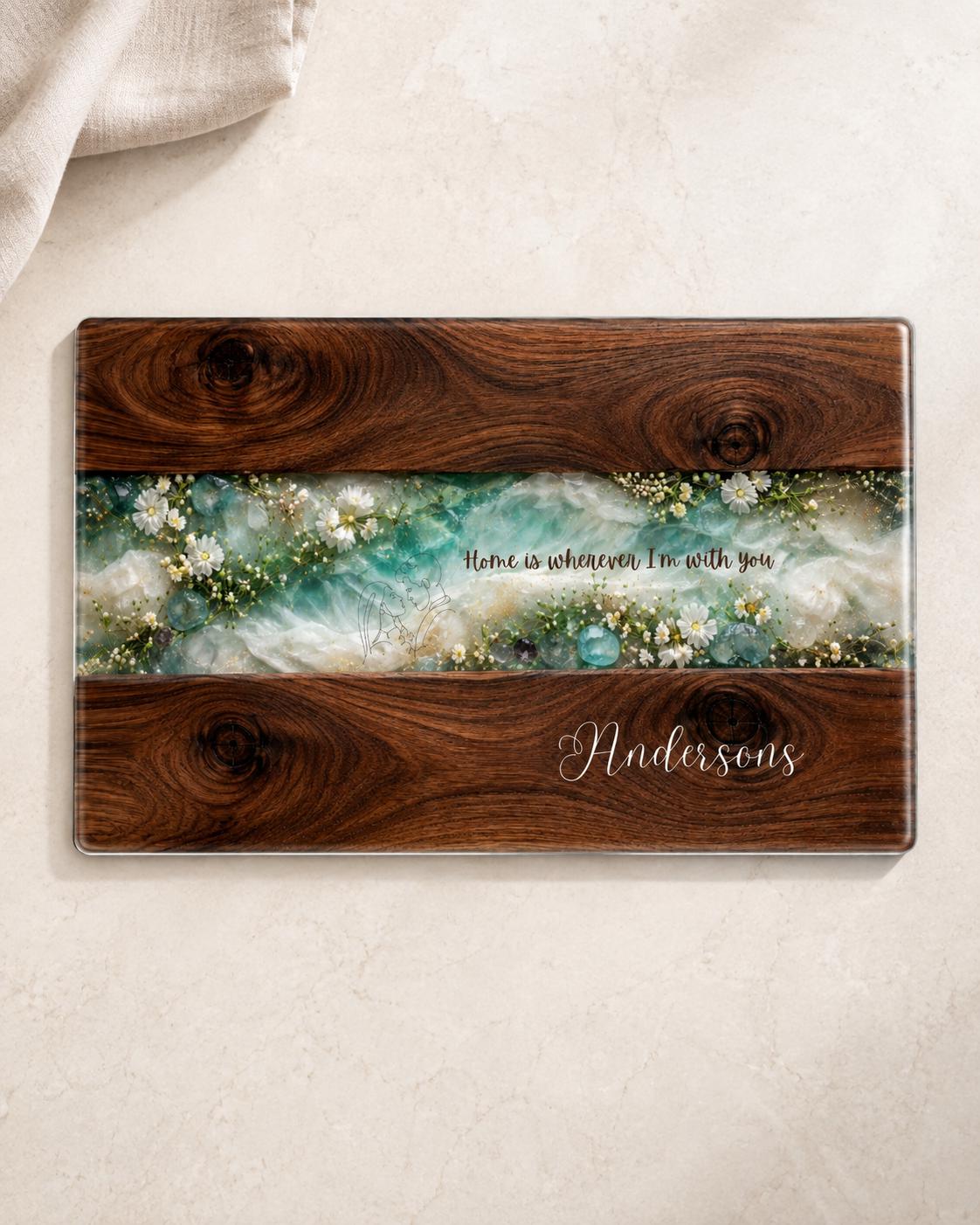 Top view of personalised glass chopping board with dark wood effect, turquoise resin-style floral centre, white wildflowers and custom Andersons family name, ideal as a housewarming gift.
