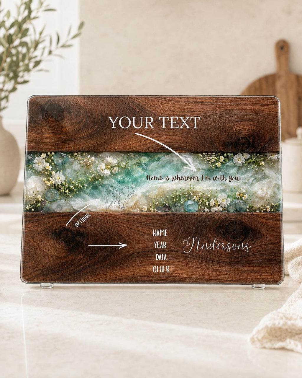 Personalised glass chopping board showing custom text options for family name, year or special message, with dark wood effect, turquoise resin-style floral centre and white wildflowers.
